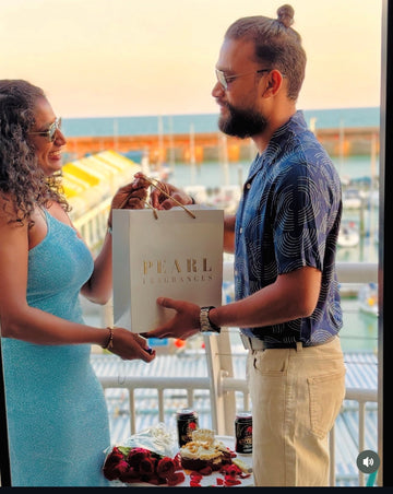 Two people standing on a balcony with a 'Pearl fragrances' bag and cake, overlooking a waterfront.